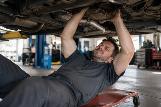 Automotive mechanic working underneath a vehicle while reaching overhead, demonstrating the physical strain that can contribute to neck and lower back discomfort, with BackFix™ promoting better posture and spinal support.